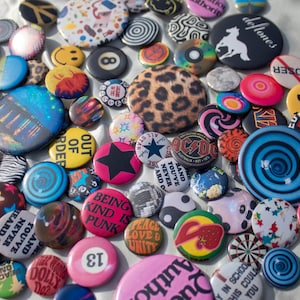 Op de afbeelding: Een verzameling kleurrijke speldjes met diverse ontwerpen, waaronder smiley gezichten, sterren en bandlogo's. De speldjes zijn in verschillende maten en vormen, met een verscheidenheid aan kleuren en patronen. Sommige speldjes hebben tekst, zoals "Being Kind is Punk".
