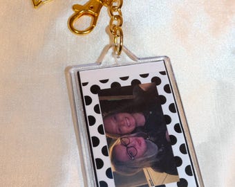 May include: A clear acrylic keychain with a photo of two people. The photo is framed with a black and white polka dot border. The keychain has a gold-colored clasp, chain, and a star charm.