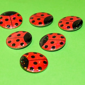 May include: Six circular cabochons featuring a red ladybird design with black spots and a glossy, glittery finish. Each piece has a rounded, domed surface and a flat back, displaying a vibrant red colour with black accents and subtle sparkles.