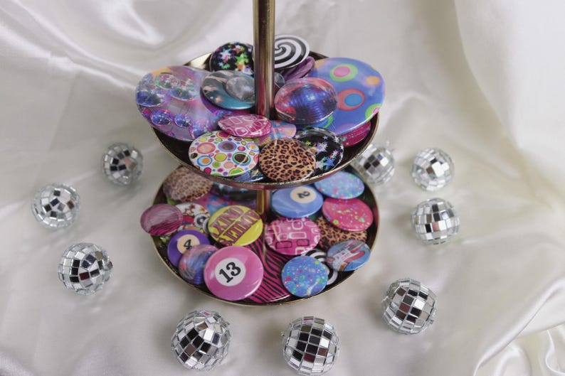 May include: A two-tiered gold-toned serving tray filled with colorful, patterned buttons. The buttons feature various designs, including galaxy, leopard print, and number 13. Silver disco balls surround the tray on a white satin surface.