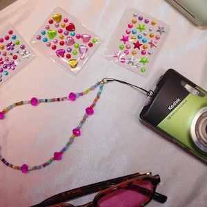 May include: A green Kodak digital camera with a black top is displayed with a beaded lanyard, colorful sticker sheets, star-shaped hair clips, and sunglasses. The camera has a silver lens and the word "Kodak" printed on it.