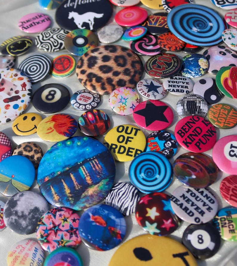 May include: A collection of colorful pinback buttons with various designs. The buttons feature images of a leopard print, a smiley face, a black star, and the text "OUT OF ORDER". Other buttons have the text "BEING KIND IS PUNK".