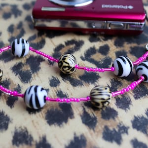 May include: A beaded bracelet with alternating zebra-striped and leopard-print beads strung on a bright pink cord. A red digital camera is in the background, resting on a leopard print surface. The bracelet is a fashion accessory.