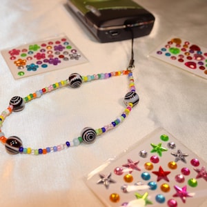 May include: A beaded phone charm with black and white striped beads and colorful smaller beads. The charm is connected to a black phone. Also visible are sheets of colorful stickers with flower, star, and gem designs.