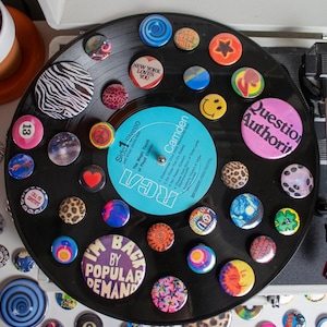May include: A collection of colorful buttons and pins adorns a black vinyl record. The buttons feature various designs, including a smiley face, a heart, and the text "Question Authority". A vintage record player is visible.