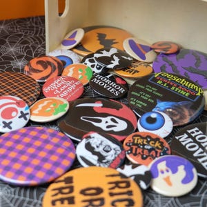 May include: A collection of Halloween-themed buttons with various designs, including pumpkins, bats, and the text "I Love Horror Movies." The buttons are in a wooden box on a spiderweb-patterned surface.