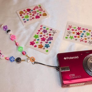 May include: A red Polaroid T730 digital camera with a beaded camera strap featuring colorful beads and charms. Three sheets of decorative stickers with hearts, flowers, and stars are also present. The camera has a silver lens and the text "7.0 Megapixels".