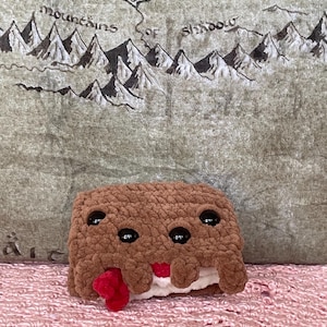 Può includere: Un giocattolo di peluche marrone a forma di barretta di cioccolato con glassa bianca e pezzi di caramelle rosse. Il giocattolo ha due occhi neri e una bocca sorridente.
