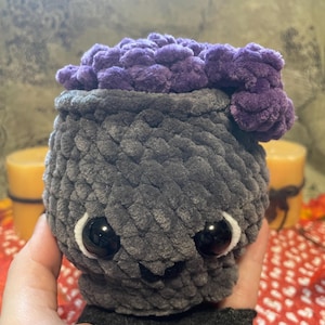 May include: A gray crocheted cauldron with a purple crocheted top. The cauldron has large black eyes and a small smile.