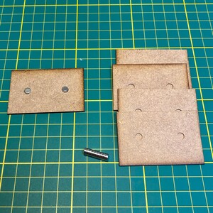 Könnte beinhalten: Ein Satz aus fünf braunen rechteckigen Holzstücken, jedes mit zwei Löchern. Die Stücke sind übereinander gestapelt. Es gibt einen kleinen Stapel silberner Magnete und ein einzelnes braunes Holzstück mit zwei Löchern auf einer grünen Schneidematte.