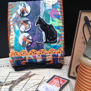 May include: A handmade mixed-media art piece featuring a black cat silhouette, decorative buttons, and orange stitching. The piece incorporates colorful paper and fabric elements, with a black ribbon and orange accents. Sewing notions and vintage ephemera are also present.