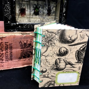 Puede incluir: Un pequeño diario hecho a mano con una cubierta marrón con un estampado botánico en blanco y negro. El diario está encuadernado con hilo verde y blanco y tiene una etiqueta en blanco en la portada.
