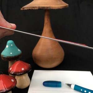 Puede incluir: Primer plano de una regla de metal plateada que est&aacute; siendo sostenida por una mano. La regla est&aacute; colocada sobre un jarr&oacute;n de madera con forma de hongo y tres peque&ntilde;os hongos pintados. Los hongos est&aacute;n pintados de rojo, azul y verde con lunares blancos.