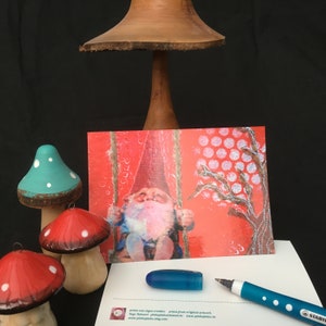 Puede incluir: Una postal roja y blanca con un gnomo en un columpio. El gnomo lleva un gorro rojo y una camisa azul. El fondo es un patr&oacute;n de lunares rojos y blancos. La postal est&aacute; sobre una superficie blanca con un bol&iacute;grafo azul y un hongo de madera.