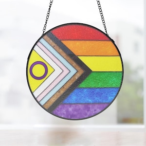 Peut inclure: Une fenêtre ronde en verre coloré suspendue à une chaîne. La fenêtre présente un drapeau arc-en-ciel de la fierté avec un cercle violet et un centre jaune. Le drapeau a un chevron noir et brun sur le côté gauche.