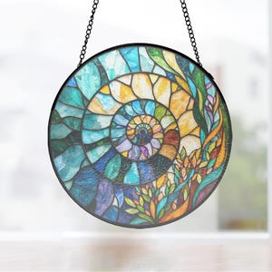 May include: A circular stained glass suncatcher featuring a spiral ammonite fossil design. The piece is composed of numerous textured glass segments in shades of blue, teal, yellow, orange, and purple, held together by dark metal cames and a black frame.