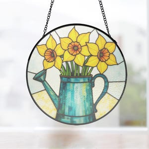 May include: Round stained glass window art featuring yellow daffodils in a teal watering can. The flowers have orange centers and green stems. The window is framed in black and hangs from a chain.