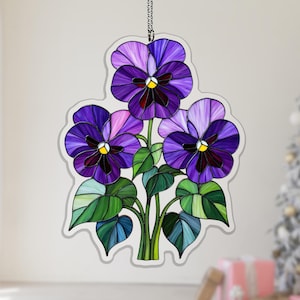 May include: A stained glass artwork depicting three purple pansies with green leaves and stems. The flowers have yellow centres and dark purple accents. The piece is outlined in a light grey border and hangs from a chain.