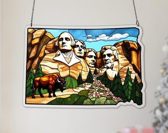 May include: Stained glass window art depicting Mount Rushmore and a bison in a landscape. The artwork is in the shape of the state of South Dakota. The window is framed with a white border and hangs from a silver chain.