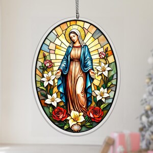 May include: Oval stained glass artwork featuring a religious figure in a blue robe, surrounded by flowers. The piece has a halo and is set against a backdrop of stained glass. The flowers are red, white, and yellow.