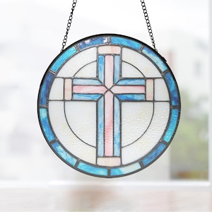 May include: Round stained glass window with a cross design. The cross is pink and blue, set against a white background. The window is framed with a dark border and a blue and white outer ring. A black chain is attached for hanging.