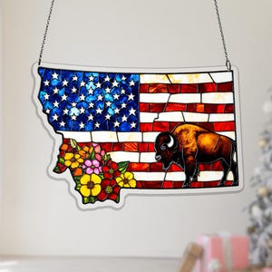 Puede incluir: Un panel de vidriera con la forma de Montana, que presenta la bandera estadounidense, un bisonte y un arreglo floral. El panel está delineado con un borde transparente y cuelga de una cadena.