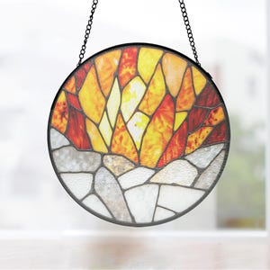 May include: Round stained glass suncatcher with a black metal frame and chain. The design features a mosaic of yellow, orange, and red glass at the top, transitioning to gray and white at the bottom. The suncatcher is hanging.