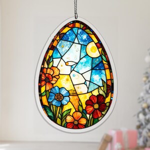 May include: An oval-shaped stained glass window hanging decoration. The design features a vibrant scene with blue skies, a yellow sun, and colorful flowers in shades of red, orange, and blue. The window is framed in white and suspended by a silver chain.