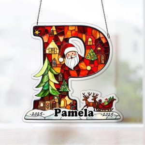 May include: A stained glass Christmas ornament in the shape of the letter "P". The ornament features Santa Claus, a sleigh with reindeer, and a village scene. The name "Pamela" is displayed, along with the year "2025".
