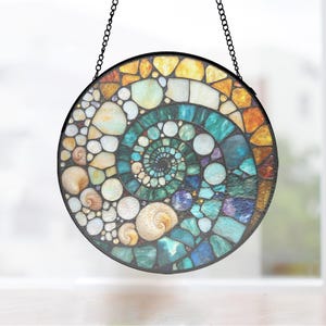 May include: A circular stained glass suncatcher featuring a spiral shell design composed of various glass pieces in shades of teal, blue, white, and amber. The artwork is framed in a thin black metal border and hangs from two black metal chains.