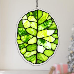May include: Stained glass leaf decoration in shades of green and yellow, with a black outline and a clear border. The leaf is suspended by a silver chain, suitable for hanging as a window or wall art piece.