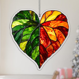 May include: Stained glass heart-shaped suncatcher with a leaf design. The left side is green and yellow, while the right side transitions from red to orange and yellow. It hangs from a silver chain.