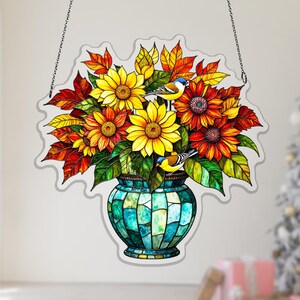 Pode incluir: Decoração de janela em vitral com um buquê de girassóis e folhas de outono em um vaso turquesa. Dois pequenos pássaros empoleiram-se entre as flores. A janela suspensa é delineada com uma corrente prateada.