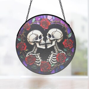Op de afbeelding: Rond gebrandschilderd glaskunstwerk met twee kussende skeletten, omringd door rode rozen en een doornige wijnstok. Het kunstwerk heeft een zwarte rand en wordt opgehangen aan een ketting. De achtergrond is een mozaïek van paars en blauw.