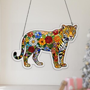 Puede incluir: Un jaguar de vidriera con un diseño floral. El jaguar se representa en una pose de pie, con su cuerpo lleno de flores coloridas. El arte en vidrio está delineado en negro y cuelga de una cadena.