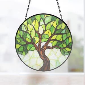 Puede incluir: Vidriera redonda con diseño de árbol. El árbol tiene un tronco y ramas marrones con hojas verdes y amarillas. El vidrio está enmarcado en negro y cuelga de una cadena negra.