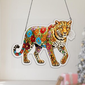Puede incluir: Un leopardo decorativo con un diseño floral. El leopardo se representa caminando, con el cuerpo cubierto de flores coloridas. La imagen cuelga de una cadena y el fondo es de color gris claro.