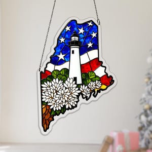 Op de afbeelding: Gebrandschilderd glaskunstwerk in de vorm van de staat Maine, met een witte vuurtoren, blauwe en witte sterren en rode strepen. Witte bloemen en groen gebladerte bevinden zich aan de onderkant. Een ketting is bevestigd om op te hangen.