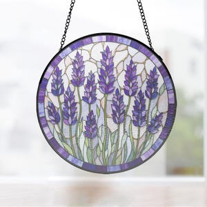 以下が含まれることがあります： 紫色の花の穂と緑の茎が描かれたラベンダー畑のデザインの円形ステンドグラスサンキャッチャー。黒い金属製の枠で縁取られ、明るい背景に対して黒い金属製のチェーンで吊り下げられています。