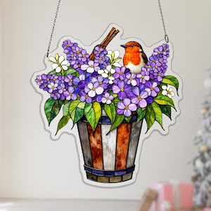 Könnte beinhalten: Dekorative Glasmalerei mit einem floralen Arrangement und einem kleinen Vogel. Das Design umfasst lila und weiße Blumen, grüne Blätter und einen braunen Vogel, der auf einem gestreiften Eimer sitzt.