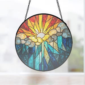 May include: Circular stained glass suncatcher with a black metal frame and hanging chain. The design depicts a sun with yellow and orange rays, clouds, and a mountain scene in blue, green, and gold hues.