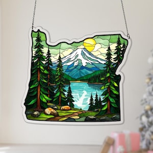 May include: Stained glass art piece in the shape of the state of Oregon, depicting a mountain landscape with a lake, trees, and a setting sun. The artwork is framed with a black outline and hangs from a chain.