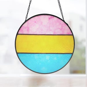 May include: A round stained glass window hanging with a black metal frame and chain. The window features three horizontal stripes: pink at the top, yellow in the middle, and blue at the bottom. The glass has a textured appearance.