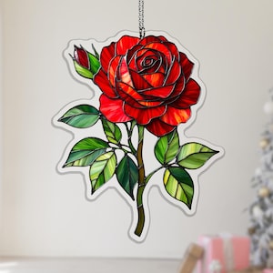 May include: A decorative stained glass-style rose. The image shows a large red rose with green leaves and a bud. The rose is detailed with shades of red and orange, and the leaves have green and yellow accents. It hangs from a silver chain.