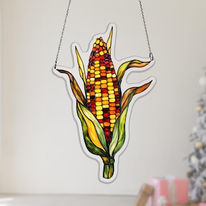 May include: A stained glass suncatcher shaped like an ear of corn with a multi-colored kernel pattern in shades of red, orange, and yellow. The corn is surrounded by green and yellow husks, outlined in black, and suspended by a thin metal chain.