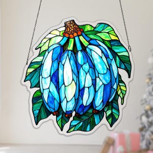 May include: A stained glass suncatcher featuring a cluster of blue bananas with green leaves and a textured orange stem. The piece is constructed from various shades of blue, green, and orange glass segments held together by dark metal solder lines.