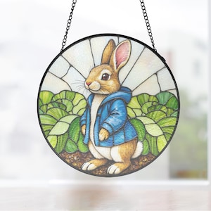 May include: A circular stained glass panel depicting a brown rabbit in a blue jacket, surrounded by green plants and a white sky. The artwork is framed in black and hangs from a chain, ideal for decorative purposes.