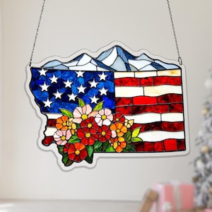 Puede incluir: Vidriera que representa la bandera estadounidense con un arreglo floral. El diseño de la bandera incorpora la forma de un estado, con estrellas azules y blancas, rayas rojas y blancas y una cadena para colgar. Un ramo de flores coloridas en la parte inferior.