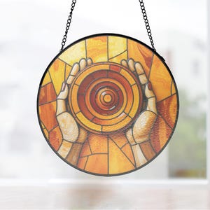 May include: Round stained glass window art featuring a design of hands cupping a circular object. The artwork uses warm tones of orange, yellow, and brown. The piece is framed in black and hangs from a chain.