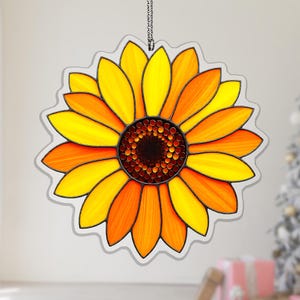 May include: A sunflower-shaped hanging ornament featuring vibrant yellow and orange petals with a dark brown, textured center. The design is outlined in black, mimicking a stained glass aesthetic, and includes a small metal chain for hanging.